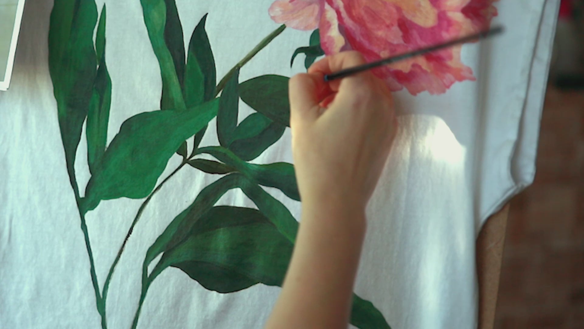 Load video: An artist painting on a white t-shirt Peony Flower. Different shots, close of of painting and the final reveal of three painted t-shirts with the same Peony motif.